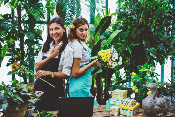 Positive young woman gardeners in aprons smiling and looking at camera while working back to back and arranging bouquets of fresh flowers - Photo, image