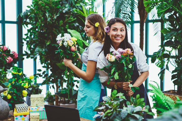 Optimistic young diverse saleswomen in aprons having fun while preparing flower compositions during work day in hothouse against green trees - Fotoğraf, Görsel