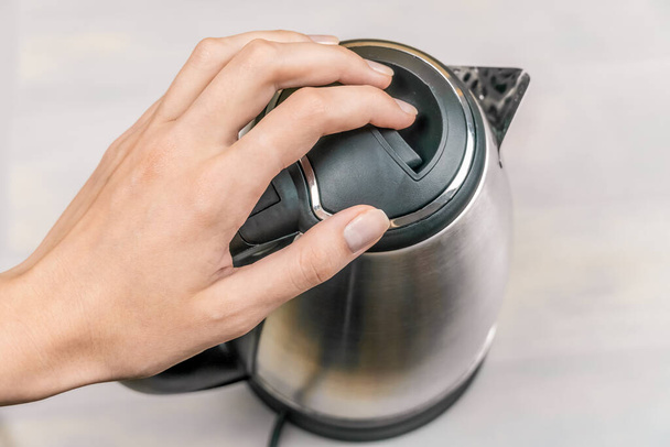 woman's hand is holding the black lid of a gray electric kettle on a light table - Φωτογραφία, εικόνα