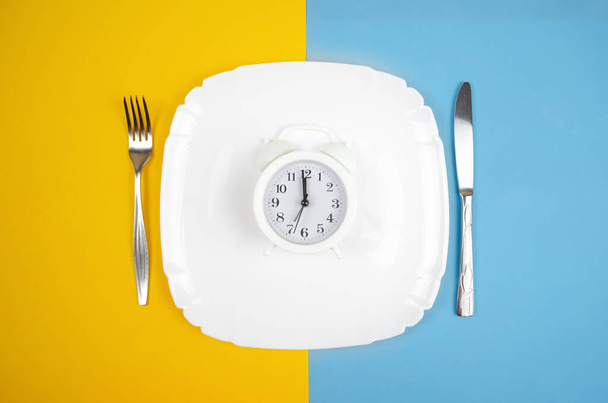 λευκό ξυπνητήρι βρίσκεται σε ένα λευκό πιάτο με ένα πιρούνι και μαχαίρι στις πλευρές σε ένα μπλε-κίτρινο φόντο, πάνω όψη - Φωτογραφία, εικόνα