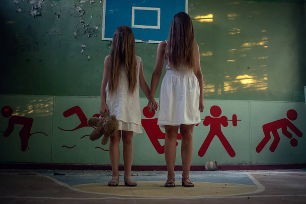 Δύο κορίτσια με άσπρα φορέματα με μακριά μαλλιά στέκονται στο εγκαταλελειμμένο γυμναστήριο ενός κατεστραμμένου σχολείου και κρατιούνται χέρι-χέρι, ένα κορίτσι που κρατάει ένα αρκουδάκι. έννοια του τρόμου, μυστικισμός - Φωτογραφία, εικόνα
