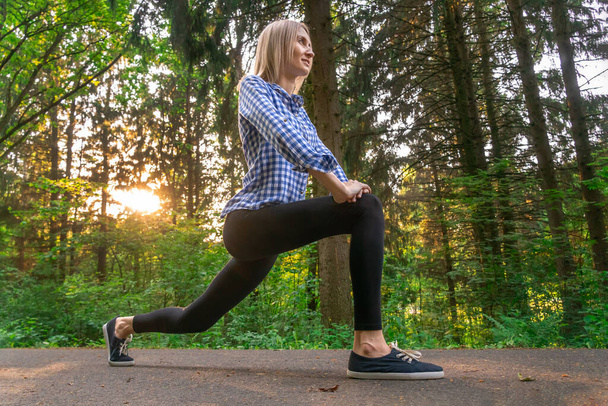 jovem bela mulher loira está envolvida em exercícios na floresta ao pôr do sol, fazendo uma lunge com os pés para a frente. - Foto, Imagem
