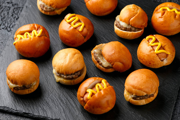 Mini hamburguesas, hamburguesas y perritos calientes con mostaza amarilla en pizarra negra - Foto, imagen