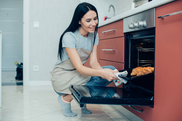 Cheerful long haired lady squatting down near the open oven and taking a baking pan with croissants out of it - Photo, image