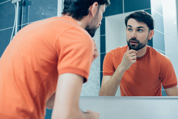 Calma hombre caucásico tocando su barba y mirando el reflejo de su cara en el espejo del baño - Foto, imagen