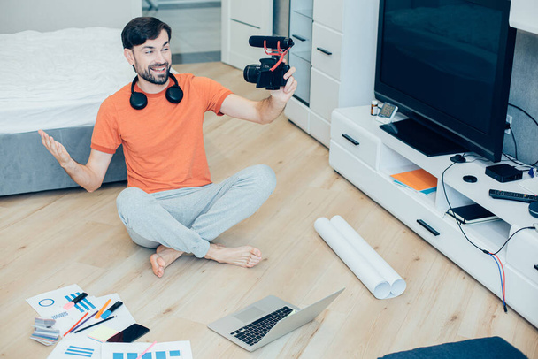 Mirthful Caucasian male blogger sitting on the floor of a bedroom and smiling while gesturing and recording himself - Photo, image