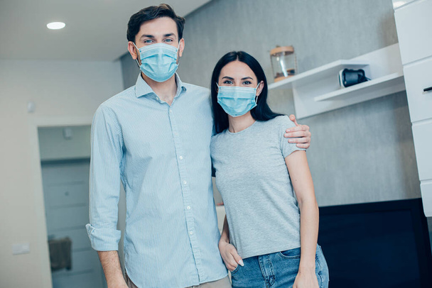 Handsome man hugging his girlfriend by her shoulder in the living room. Medical masks on their faces - Photo, image