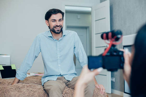 Mirthful handsome man sitting on a bed at home and smiling to the camera with a woman recording him - Фото, зображення