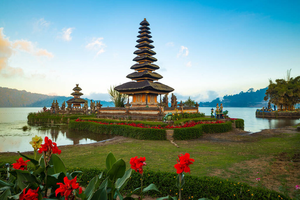 Pura Ulun Danu Bratan tempel op Bali eiland. Prachtige balinese tempel. Balinese mijlpaal. Bewolkte lucht. Waterreflectie. Voorgrond met rode bloemen. Bratan Lake, Bedugul, Bali, Indonesië - Foto, afbeelding