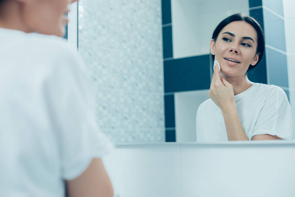 Calm beautiful woman looking at the reflection in the bathroom mirror while cleaning her facial skin with a cotton pad - Photo, Image