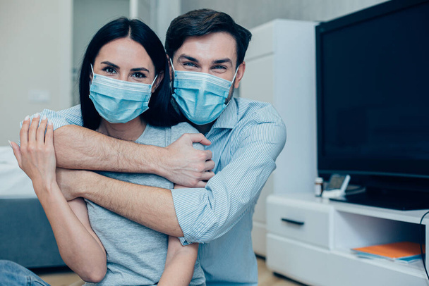 Handsome man sitting in the room behind his beautiful girlfriend and embracing her. Medical masks on the faces - Fotoğraf, Görsel