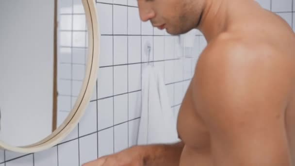 hombre sin camisa sosteniendo contenedor y aplicando crema facial en el baño - Imágenes, Vídeo