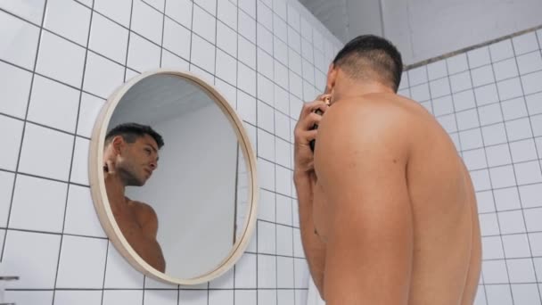 vista basso angolo di uomo senza camicia spruzzando profumo e guardando lo specchio - Filmati, video