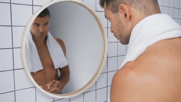 Мужчина без рубашки наносит лосьон после бритья и смотрит в зеркало  - Кадры, видео