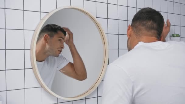 jovem em branco t-shirt fixando o cabelo e olhando para o espelho - Filmagem, Vídeo