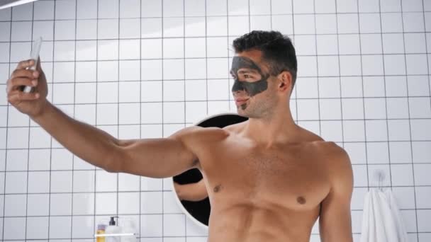 hombre sin camisa con máscara de arcilla en la cara tomando selfie en el baño - Imágenes, Vídeo