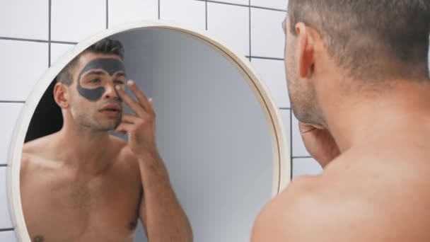όμορφος άντρας με μάσκα από πηλό στο πρόσωπο και κοιτώντας τον καθρέφτη - Πλάνα, βίντεο