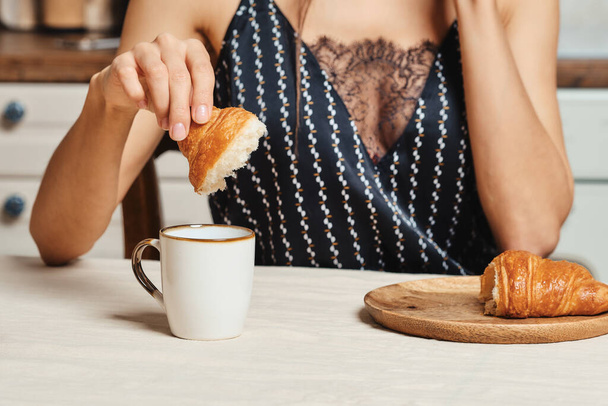 Hermosa mujer de raza mixta comiendo croissant fresco en la cocina por la mañana - Foto, Imagen