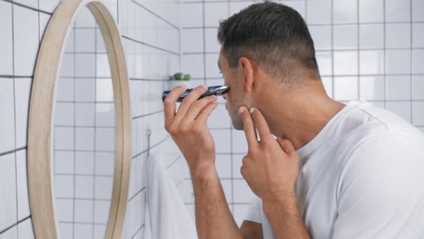 άνδρας σε λευκό t-shirt χρησιμοποιώντας trimmer, ενώ το ξύρισμα πρόσωπο στο μπάνιο  - Πλάνα, βίντεο