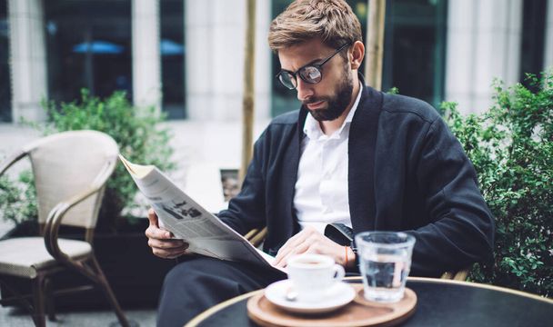 Serious bearded well dressed businessman in eyeglasses reading last news in newspaper while sitting with cup of coffee in street cafe - Foto, imagen