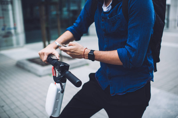Crop faceless male wearing casual denim shirt wristwatch and black backpack browsing mobile phone on scooter on pavement during daytime - 写真・画像