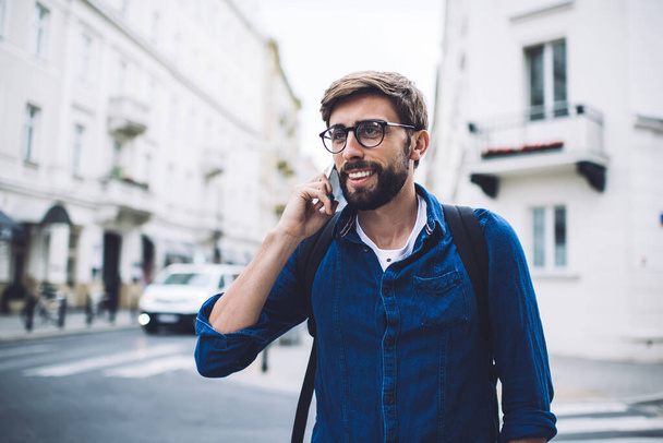 Satisfied bearded youthful hipster wearing casual denim shirt and eyeglasses talking on mobile phone while crossing road and looking away - Fotografie, Obrázek