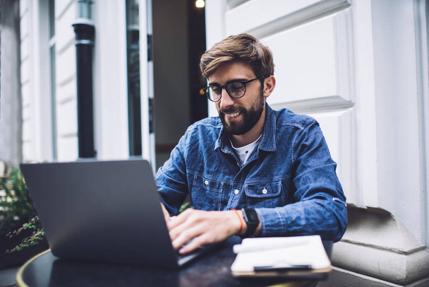 Adult bearded smiling remote worker in eyeglasses and casual outfit with smart watch typing on netbook while sitting at round table on street in afternoon - 写真・画像