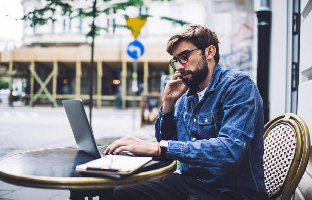 Side view of male remote worker in denim jacket speaking on cellphone and typing on netbook while sitting at black table on street in afternoon - Φωτογραφία, εικόνα
