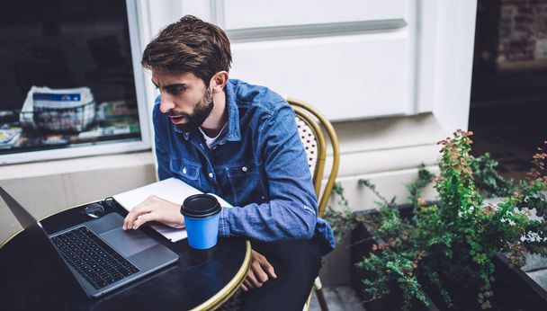 Focused young man in casual outfit concentrating on screen of laptop and immersing in working process with cup of coffee to go on table - Fotó, kép