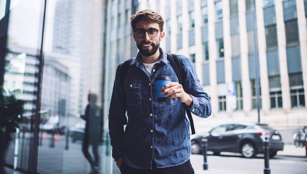 Confident bearded male wearing casual clothes and eyeglasses standing with coffee and looking at camera while drinking near buildings in modern city - Valokuva, kuva