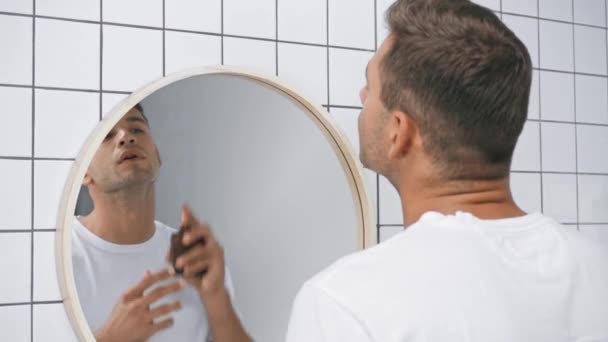 mies valkoisessa t-paidassa ruiskuttamalla hajuvettä ja katsomalla peiliin  - Materiaali, video