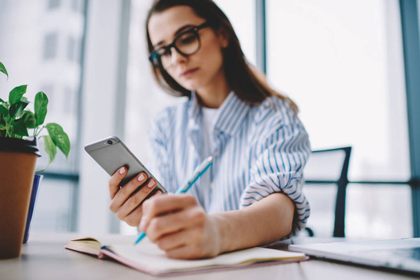 Millennial female student sitting at table desktop with modern smartphone gadget and textbook planner for e learning, blurred woman using mobile phone for informative research while making notes - Fotografie, Obrázek