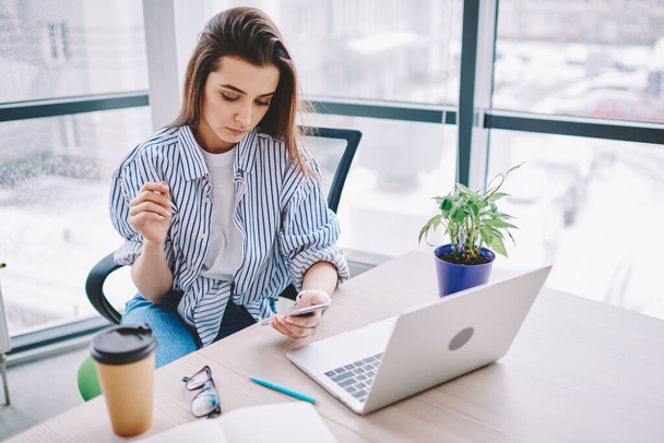 Caucasian female freelancer checking mobile notification during remote job in modern workspace, skilled millennial in smart casual wear making online booking and banking via smartphone application - Foto, Imagem