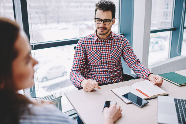 Caucasian male employee in eyewear for vision correction listening attentively blurred female colleague talking about together startup projecting, smart casual partners planning business strategy - Foto, afbeelding