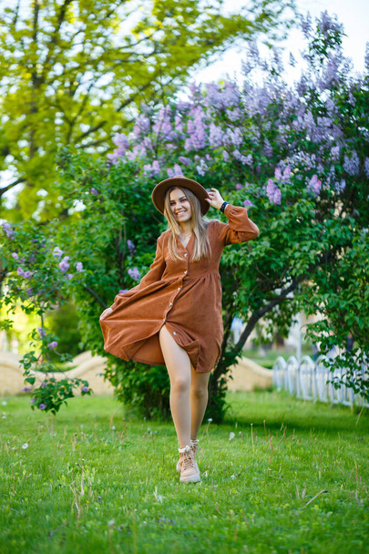 Uma menina de chapéu marrom e vestido caminha pelo parque, ela tem uma aparência europeia e um sorriso em seu rosto. Jovem mulher atraente caminha em um dia ensolarado - Foto, Imagem