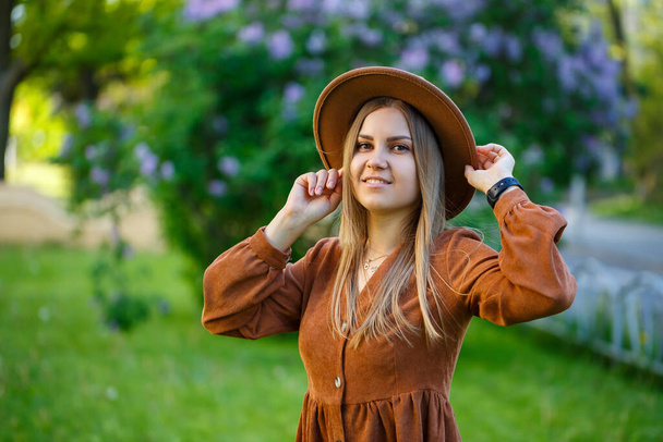 Молода стильна дівчина в капелюсі біля пишного куща рожевого бузку в саду. Дівчина насолоджується запахом бузку. Теплий літній день в саду молода жінка з посмішкою на обличчі
 - Фото, зображення