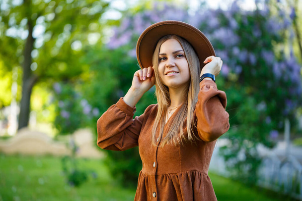 Красивая девушка с светлыми волосами держит шляпу в руках и стоит с закрытыми глазами рядом с лиловым кустом. Молодая женщина в саду с цветущими деревьями - Фото, изображение