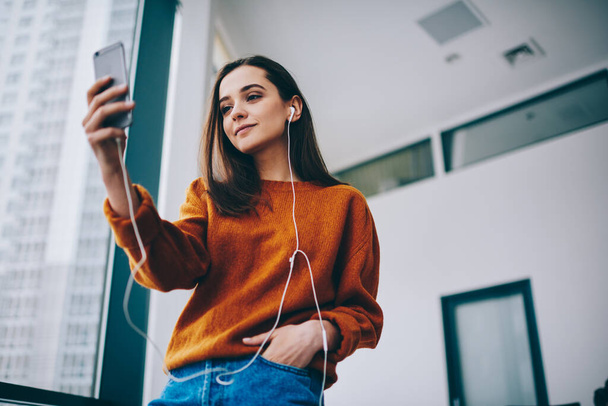 Caucasian hipster girl taking pictures during leisure time using front camera on smartphone gadget enjoying favourite soundtrack, attractive female millennial listening music playlist indoors - Foto, Imagem