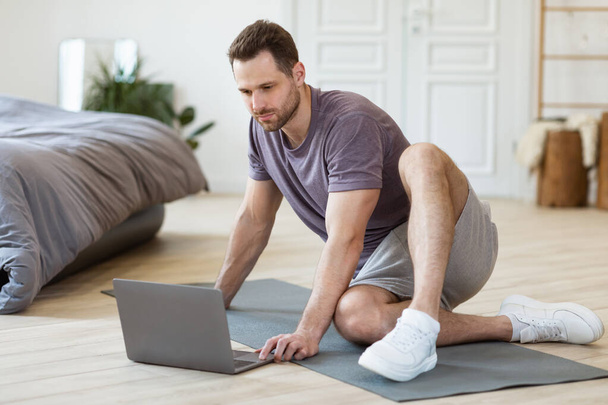 Homem com laptop sentado no tapete pronto para o treinamento dentro de casa - Foto, Imagem