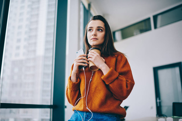 Pensive hipster girl in stereo headphones holding disposable takeaway cup listening book and thinking about audio information, contemplative female millennial in earphones pondering on song text - Foto, imagen