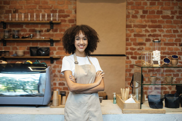 Sonriente joven afroamericana dama en delantal se para cerca del mostrador de bar con equipo para hacer café y mira a la cámara - Foto, Imagen