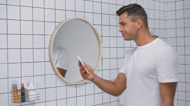 hombre alegre en camiseta blanca tomando selfie en teléfono inteligente en el baño  - Metraje, vídeo
