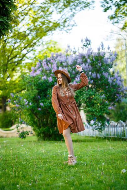 stijlvol meisje met een bruine hoed en een lichte jurk op een achtergrond van lila weelderige struiken. Jonge vrouw met een glimlach op haar gezicht op een zonnige zomerse wandeling in het park - Foto, afbeelding