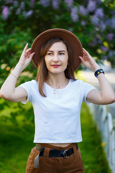 Πορτρέτο της ελκυστική κομψή γυναίκα σε ένα καπέλο με μακριά ξανθά μαλλιά στο πάρκο, ντυμένος με ένα λευκό t-shirt και καφέ παντελόνι. - Φωτογραφία, εικόνα