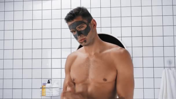 mężczyzna bez koszuli z glinianą maską na twarzy, naprawiający włosy patrząc w kamerę - Materiał filmowy, wideo