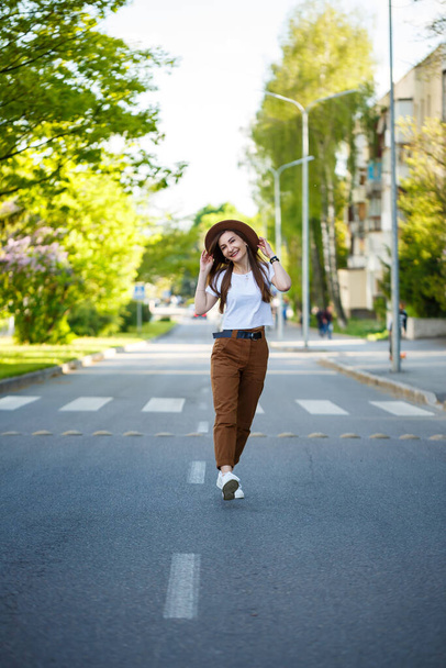 Een mooi meisje met een bruine hoed en een wit T-shirt loopt op een warme zonnige dag over de weg. mooie jonge vrouw in een hoed met een glimlach op haar gezicht - Foto, afbeelding