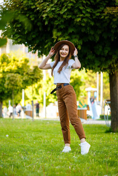 Young happy woman in a hat walks along an alley in a park. A girl of European appearance with a smile on her face on a bright sunny summer day - Foto, Bild