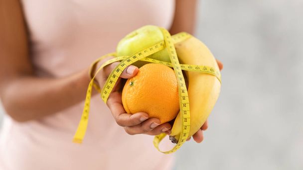 Healthy diet for weight loss concept. Closeup view of black lady holding fresh fruits with measuring tape - Foto, imagen