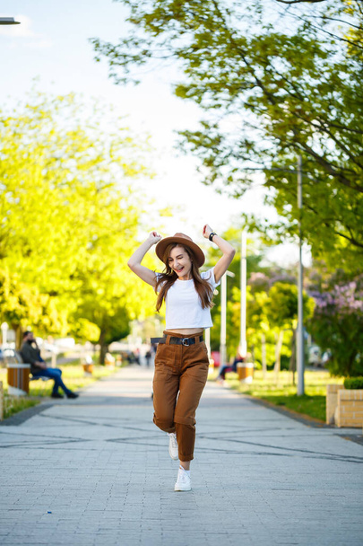 Молодая счастливая женщина в шляпе ходит по аллее в парке. Девушка европейского вида с улыбкой на лице в яркий солнечный летний день - Фото, изображение