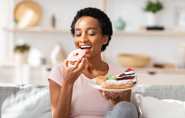 Молода чорна жінка на дієті зраджує день їжі, тримає тарілку цукерок, фаршируючи рот пончиком вдома
 - Фото, зображення